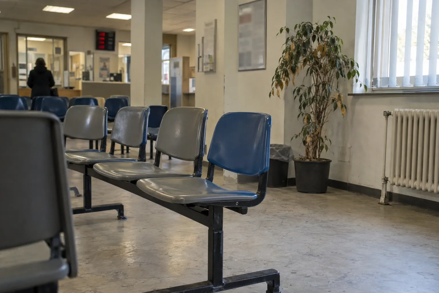 Зал ожидания в немецком Jobcenter: ряды пластиковых стульев, фикус и табло очереди на заднем плане