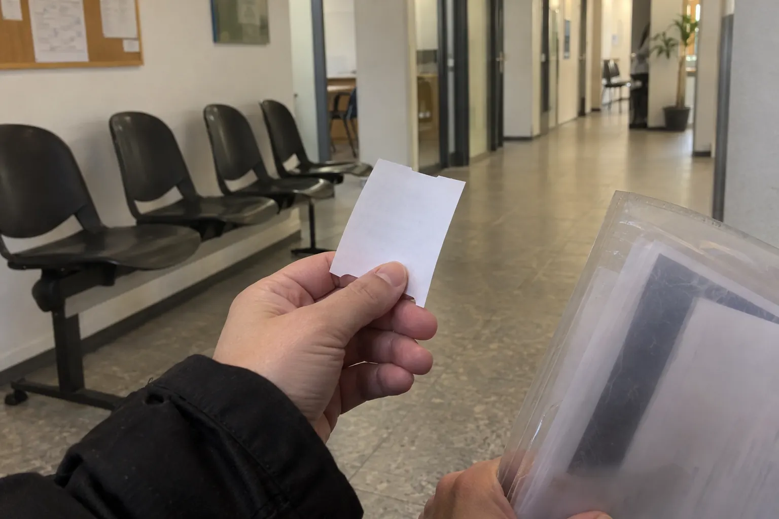 Рука держит талон электронной очереди, в другой руке прозрачная папка с документами, сзади стулья в коридоре